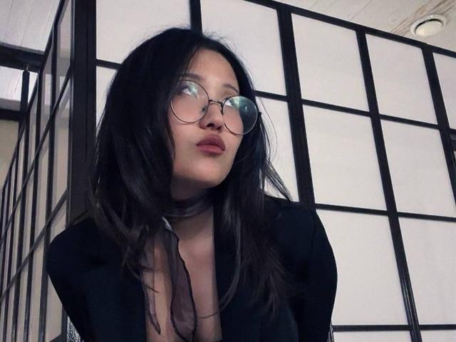 Alvina Njie, avec ses longs cheveux noirs, portant des lunettes rondes, un collier noir et une tenue noire moulante qui dévoile sa poitrine, se tient devant un écran shoji de style japonais..