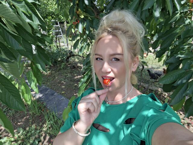 Blond MerlynonYou with green top, holding red apple, biting it, smiling, wearing necklace and bracelet, standing under leafy tree, sunlight filtering through.