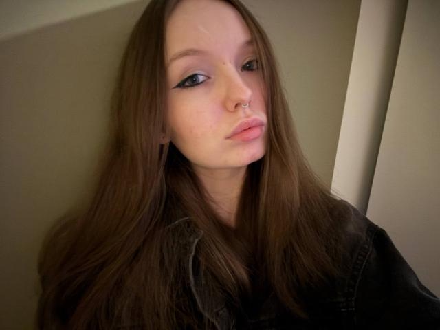 NieshaRonsini has long brown hair, a nose ring, black eyeliner, and is pouting with pink lips, wearing a black denim jacket. Her skin is pale and she looks sexy.