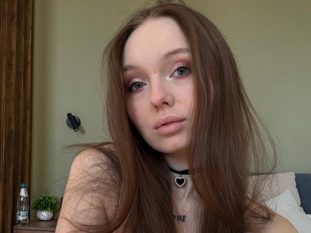 NieshaRonsini has long brown hair, pink eye makeup, a nose ring, and a black choker with a heart pendant. She's looking at the camera with pale skin.