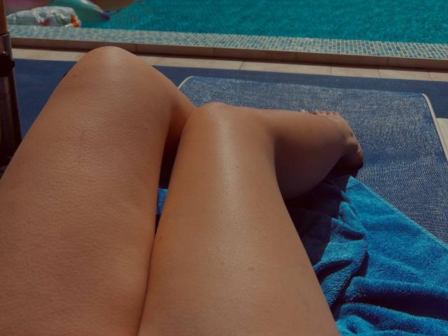 AlexXas' sun-kissed thighs and smooth, tan skin are on a blue towel by a pool, with their toes touching the water. Their legs are glistening with water drops.