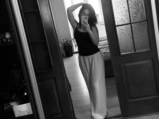 Black-and-white selfie of AlexXas with arms up, wearing a black tank top and high-waisted, pleated white pants, standing in front of a door, holding a phone.