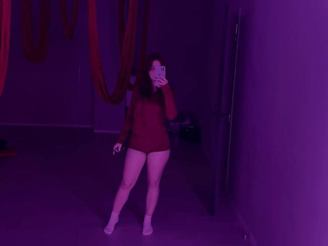 AlexXas with red long-sleeve leotard and white socks, holding phone, purple-lit room, dark red drapes, standing, legs apart, visible pussy, medium brown curly hair, covering face.