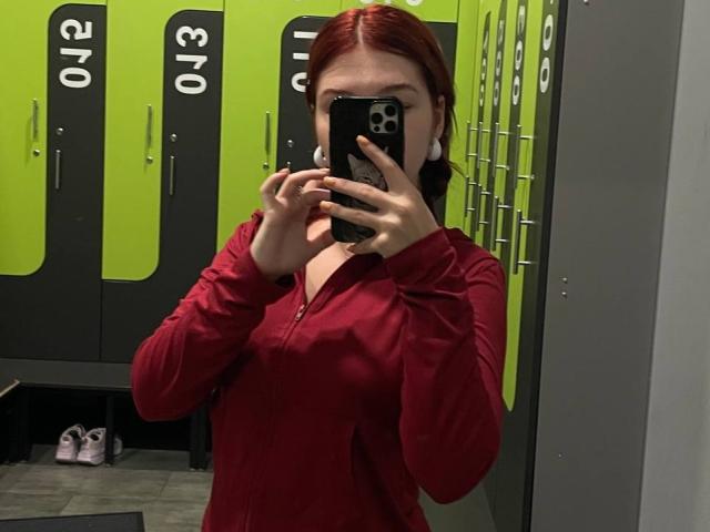 AlexXas with red hair, wearing a red zip-up, holding a phone covering her face, taking a mirror selfie in a locker room. Boobs are slightly visible through the shirt.