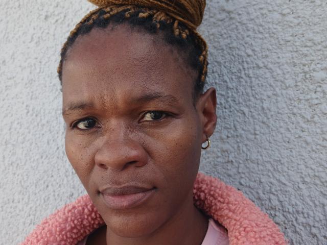 SassyQueen has dark brown skin, small gold hoop earring, braided hair in a bun, pink textured jacket, and a serious expression. Her eyes are slightly green and she's looking at camera.