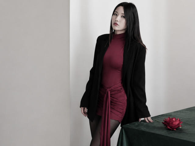 Alvina Njie, con cabello negro largo, vestida con un vestido rojo ajustado, una chaqueta negra y medias negras, está de pie junto a una mesa verde con una flor roja. Se puede ver su trasero..