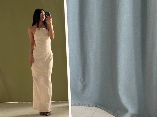 AlvinaNjie standing in a full-length mirror, wearing a white, sleeveless, lace dress, holding a phone, with long black hair, green background, and blue curtain on the right.