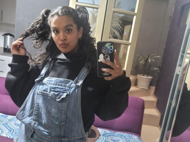 ZiaNoir, avec ses cheveux noirs bouclés attachés en queue de cheval, portant un pull noir à col roulé et une salopette en jean bleu, prend un selfie devant un miroir, tient ses cheveux et regarde l'appareil photo..
