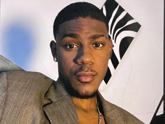 DrakeWit with short black hair, brown skin, small goatee, wearing a brown pinstripe jacket and silver necklace, looking slightly to the side with a neutral expression.