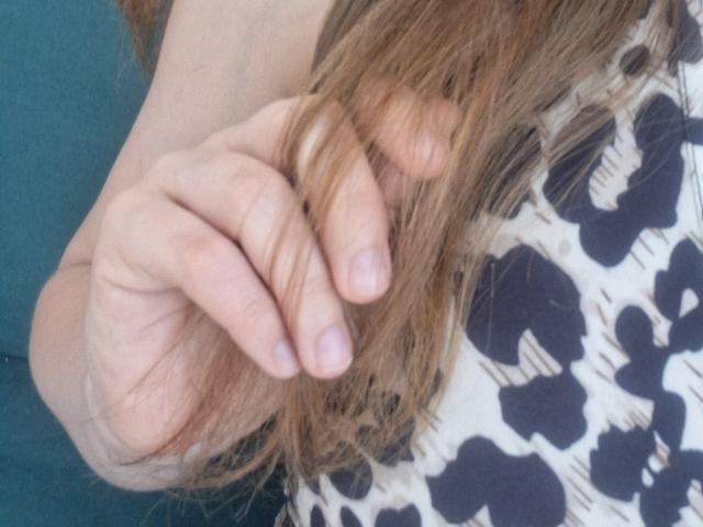 La mano de Marriam, con cabello corto, castaño y ligeramente ondulado, sostiene suavemente una sección de su cabello. Lleva una blusa con un estampado en blanco y negro. Sus uñas están limpias..