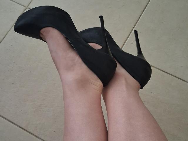 Las piernas desnudas de Marriam, con tacones altos negros de punta, con los pies cruzados a la altura de los tobillos, mostrando las plantas y la parte inferior de los tacones..