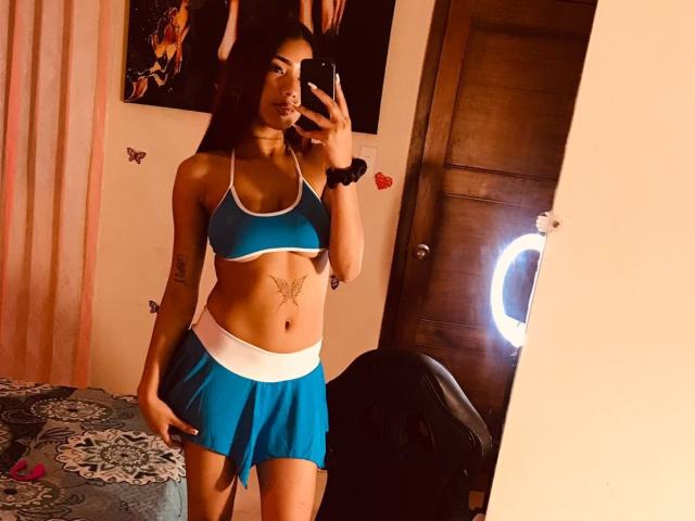 DakotaBrenda con un top deportivo y falda azules, mostrando sus tatuajes, incluyendo una mariposa en la parte baja del abdomen, posando en un selfie frente a un espejo, sosteniendo un teléfono..