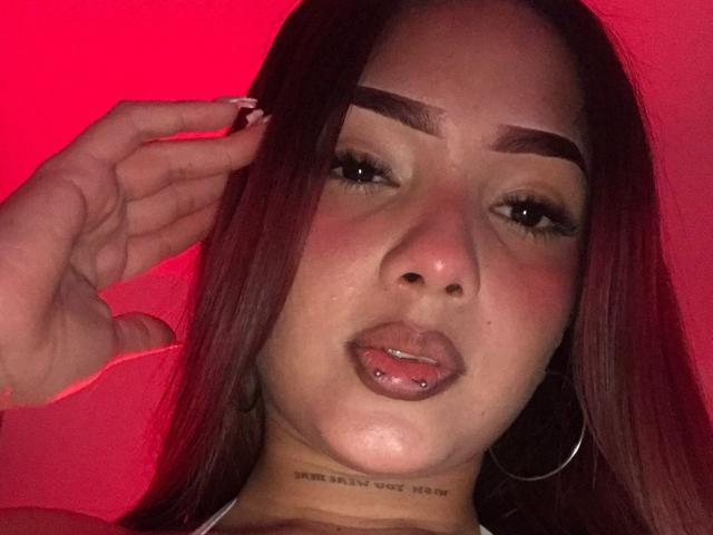 DakotaBrenda with red-painted nails, full lips with a piercing, dark eyebrows, long brown hair, and hoop earrings, against a red-lit background, looking slightly annoyed.