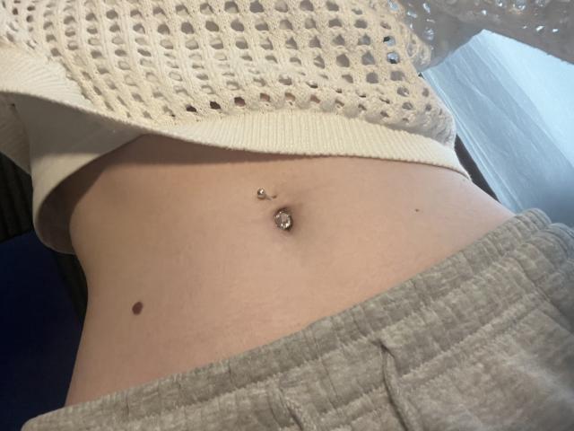 Ginnia's midriff with a pierced navel and a small brown mole, wearing a white knit top and gray pants, in a slightly angled, close-up shot.
