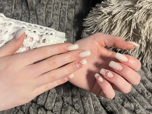 Le mani di Ginnia, con la pelle chiara, le unghie lunghe color beige, e la decorazione floreale rossa, riposano su una coperta grigia con una texture particolare. Sullo sfondo, un cuscino bianco all'uncinetto e un cuscino beige con frange..