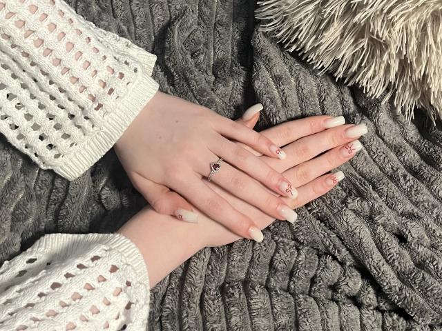 Le mani di Ginnia, con unghie lunghe, bianche e appuntite, decorate con motivi floreali rosa e con un anello, poggiate su una coperta grigia, testurizzata e lavorata a maglia, indossa una manica bianca all'uncinetto..