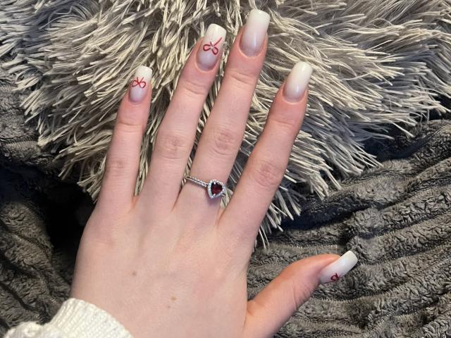 Ginnia's hand with white nails, red bow designs, and a heart ring with a red gem on a fluffy gray blanket. Her skin is pale, and she wears a white sleeve.
