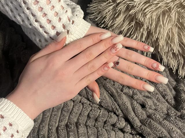 Le mani di Ginnia, con la pelle chiara, le lunghe unghie bianche in acrilico, la decorazione a forma di croce rossa sulle unghie e un anello con una pietra rossa sul mignolo, riposano su una coperta grigia con una trama particolare..
