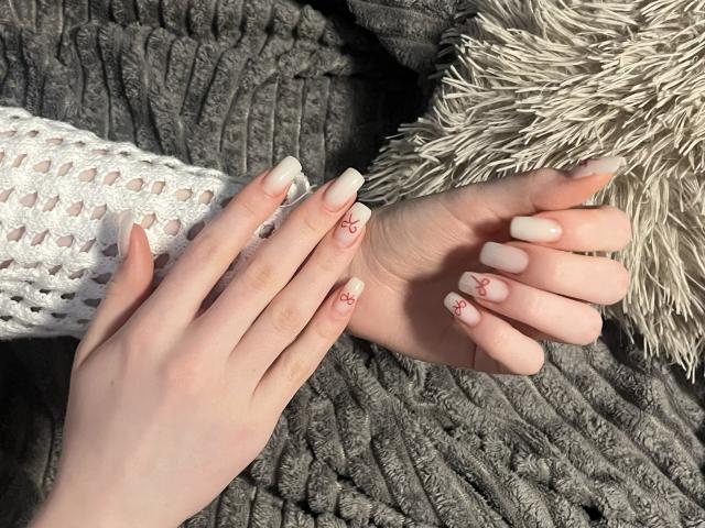 Ginnia's pale hands with white, crocheted sleeves and white, stiletto nails with red patterns, resting on a gray, textured blanket with a fluffy, beige pillow.