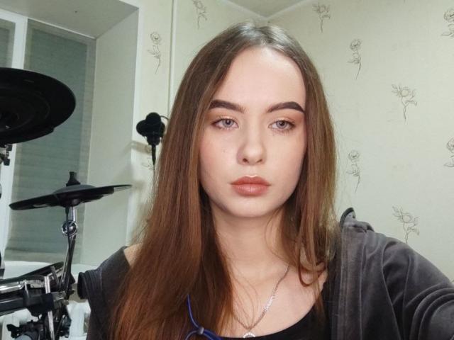 SammieFluke heeft lang bruin haar, een lichte huid en volle lippen. Ze draagt een zwarte top met een zilveren ketting. Achtergrond: drums..