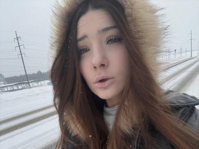 Angie Plassmeyer, con i capelli castani, indossa un cappuccio di pelliccia, sembra infastidita, ha della neve sul viso, indossa una giacca grigia, sullo sfondo si vede una strada, il cielo è nuvoloso, si vedono fiocchi di neve, le labbra sono leggermente socchiuse..