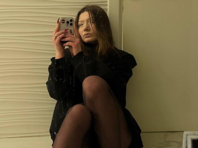 AngiePlassmeyer with wet hair, black dress, and sheer black stockings, holding a phone, legs crossed, serious expression, reflective mirror, water droplets.