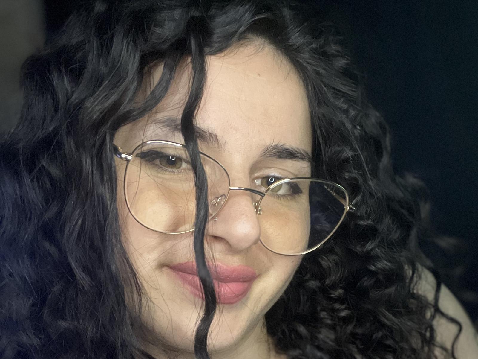 NaughtyRoseOne tiene el pelo negro y rizado, usa gafas redondas y tiene pintalabios rosa, con una ligera sonrisa, mostrando una piel clara y ojos marrones. Su nariz es pequeña y linda..
