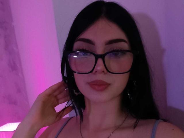IsabellaaRossi with black hair, glasses, and pink lipstick, touching her ear, wearing a grey top, in purple lighting. Her tits are small, she's cute.