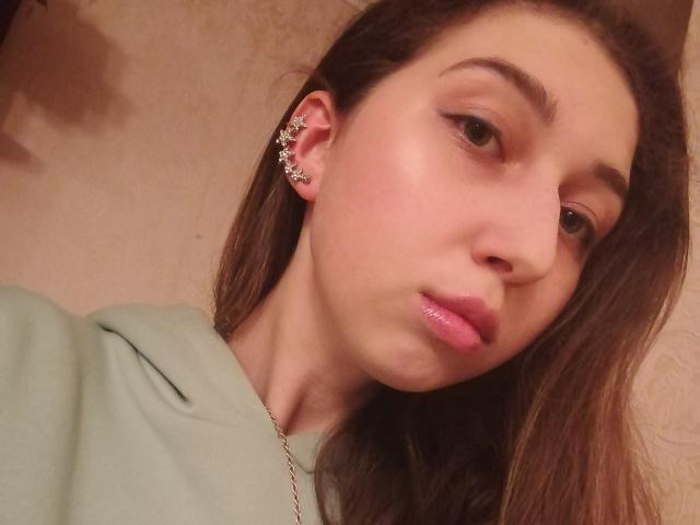 Sanella with shiny pink lips, light green shirt, brown hair, and a silver ear cuff, looking slightly to the side with a subtle pout. Soft, slightly flushed skin.