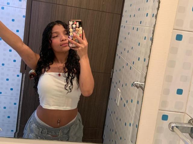 LightenStars with curly black hair, white tube top, showing her midriff, grey pants, and navel piercing, taking a mirror selfie with a phone decorated in colorful stickers.
