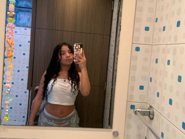Mirror selfie of LightenStars with long curly black hair, white crop top, and gray pants showing a belly button piercing, holding a phone with a colorful case.
