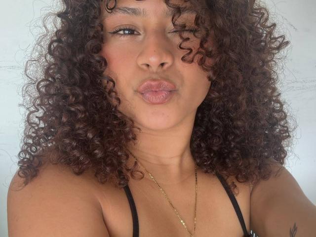 SofiaPassionne with curly brown hair, pouty lips, tan skin, wearing a black top, gold necklace, and a subtle tattoo on her right shoulder. She's making a kissing face.