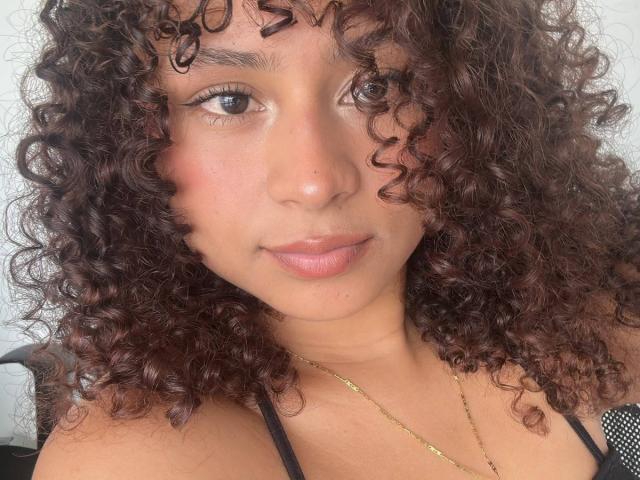SofiaPassionne with curly brown hair, light brown skin, and small nose, wearing a black top, gold necklace, and pink lipstick, staring into camera with a subtle smile.
