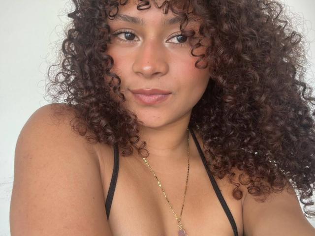 SofiaPassionne has curly brown hair, medium brown skin, and is wearing a black halter top with a gold necklace. She has small tits and a neutral expression.