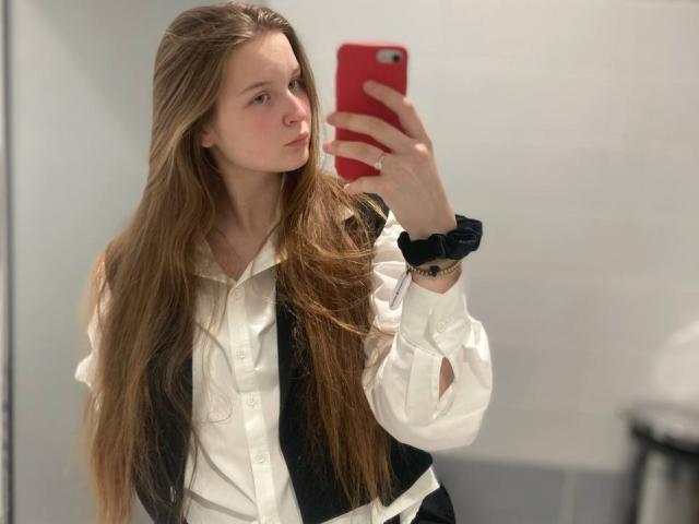 JuttaHillwig with long brown hair, white shirt, black vest, holding red phone, black scrunchie on wrist, taking mirror selfie, looking serious, showing small tits, in bathroom.