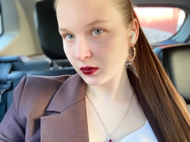 JuttaHillwig with fair skin, blue eyes, and red lipstick, wearing a brown blazer over a white top, and a silver necklace with a cross, in a car.