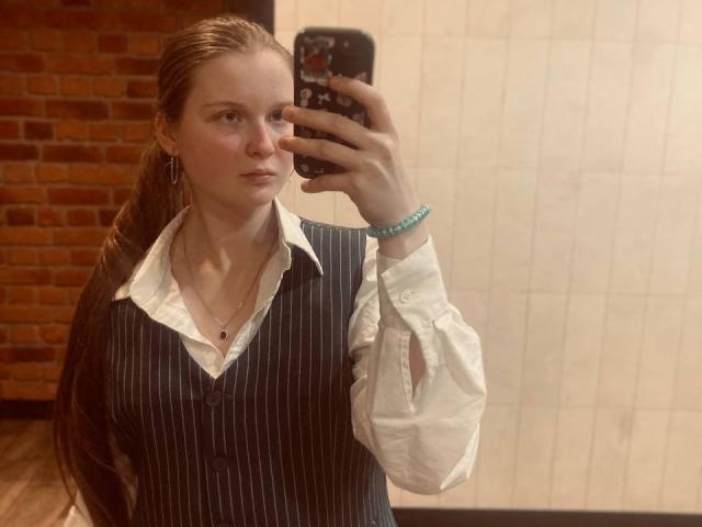 JuttaHillwig with pinstriped vest, white shirt, and bracelet, taking a mirror selfie with phone in hand, showing pale skin and brown hair, standing in a room.