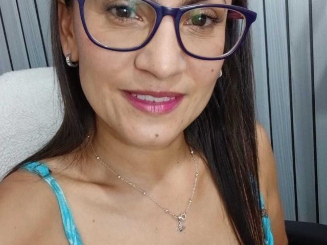 SolaraDubois with big purple glasses, pink lipstick, blue top, necklace, and earrings, showing a smile with small moles on her face, and straight brown hair.