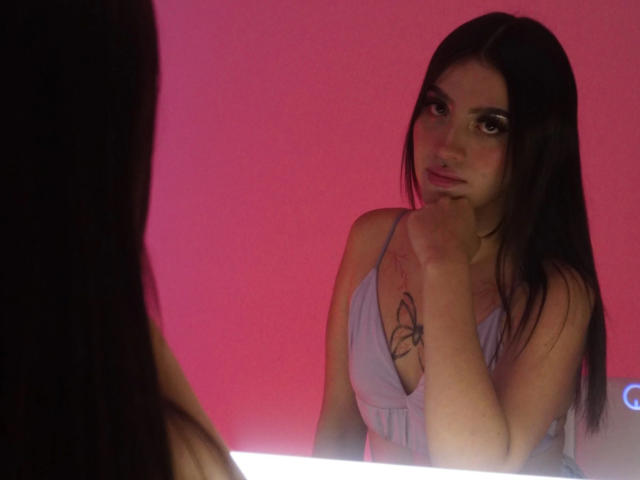 LunaaBlossomm with long dark hair, wearing a lilac top, has a butterfly tattoo on her chest, looking serious with her hand near her mouth, in a pink-lit room.