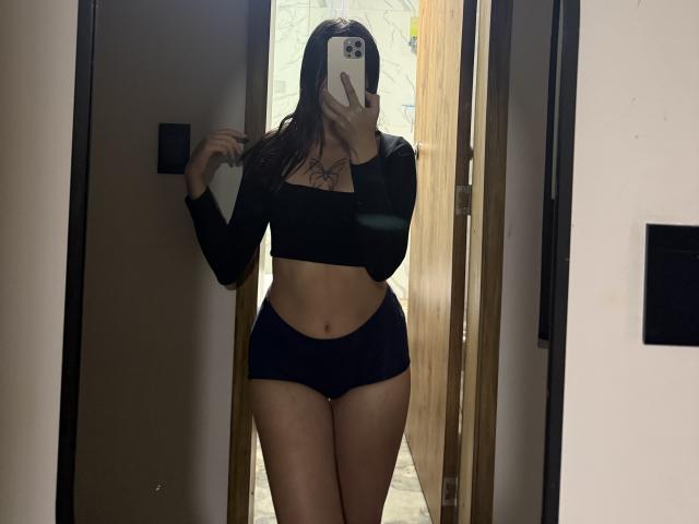 LunaaBlossomm, con i capelli scuri, indossa un top corto a maniche lunghe nero e biancheria intima blu navy, è in piedi in una porta e tiene un telefono davanti al viso. Tatuaggio visibile sul petto..