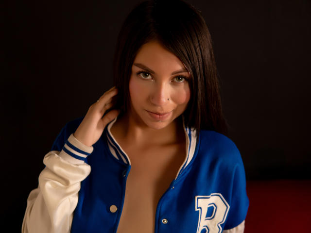 AlinnaSans with dark brown hair, wearing a blue and white varsity jacket, slightly open, exposing her bare chest and small, perky tits, looking directly at the camera.
