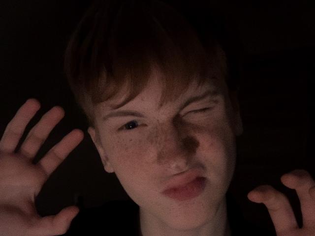 JoshuaGussy has freckled skin, winks with one eye, and makes a pouty face while raising both hands, dark background, short brown hair, and a slightly goofy expression.
