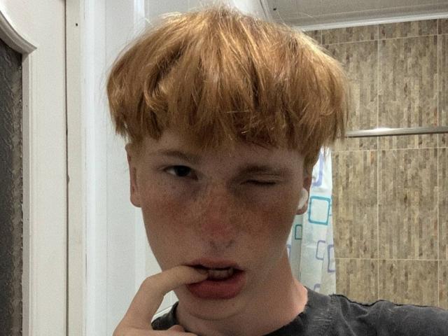 JoshuaGussy met rommelig rood haar, knippend met zijn oog, een vinger bijtend, een bezaaid gezicht, licht open mond, gekleed in een zwart shirt, in een badkamer met beige tegels..