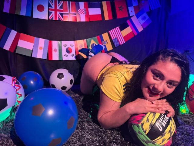 CristalZafiro knielend, geel top, zwarte string, een voetbal vasthoudend, billen omhoog, lachend, blauwe ballonnen, voetballen, vlaggen, donker haar, pluizig tapijt, blauwe verlichting..