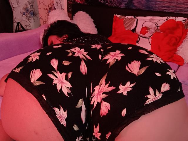 AmberSunshine, ass visible, portant un short noir à motifs floraux, allongée face contre face sur un canapé violet, avec des coussins rouges et blancs, un soutien-gorge rouge, des cheveux noirs, un éclairage rose, une silhouette pulpeuse, des fesses rebondies..