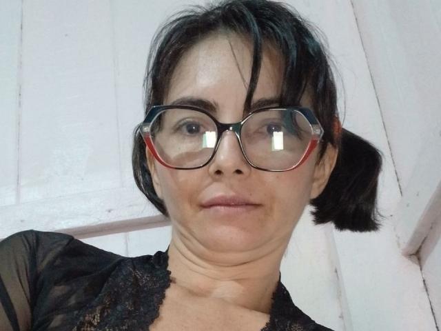 Luaunt, avec des lunettes, un haut en dentelle noire, de courtes couettes et un visage légèrement ridé, se prend en selfie en mettant ses seins en valeur. Ses lunettes à monture rouge reflètent la lumière..