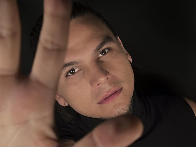 Das Gesicht von RobSaenz mit langen, dunklen Haaren, braunen Augen und heller Haut, in einem schwarzen Hemd, blickt in die Kamera, wobei seine rechte Hand im Vordergrund zu sehen ist..