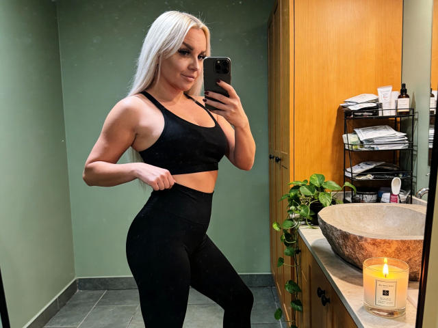 MiaXXX, une femme séduisante avec de longs cheveux blonds, un soutien-gorge de sport noir et un legging noir taille haute, tenant un téléphone, debout dans une salle de bain avec une bougie sur le comptoir..