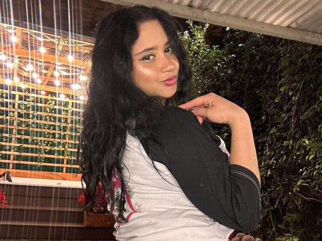 GraceBlue69 with long black wavy hair, light brown skin, and pink lipstick, wearing a black and white top, looking over her shoulder, hand near her neck.