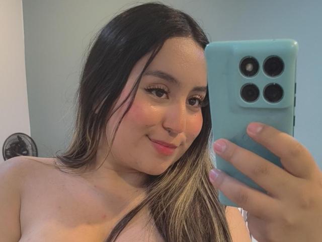 SalomettX, com cabelos longos e escuros, um telefone azul e batom rosa, segurando o telefone, mostrando os ombros, sorrindo para o espelho, com as bochechas levemente coradas, rosto fofo, nariz pequeno, sorriso discreto..